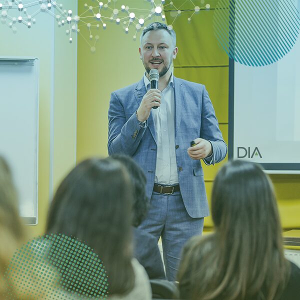 A man in a blue suit speaks into a microphone before an audience in a brightly lit room with yellow walls at an International Society for Medical Publication Professionals event.