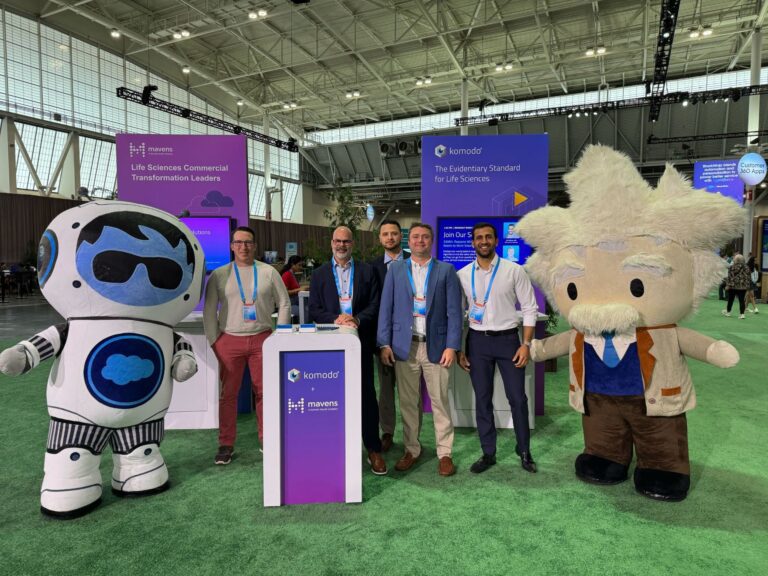 Five people, including two in robot and Einstein costumes, stand together at a Komodo Health booth at a life sciences event in a convention center.