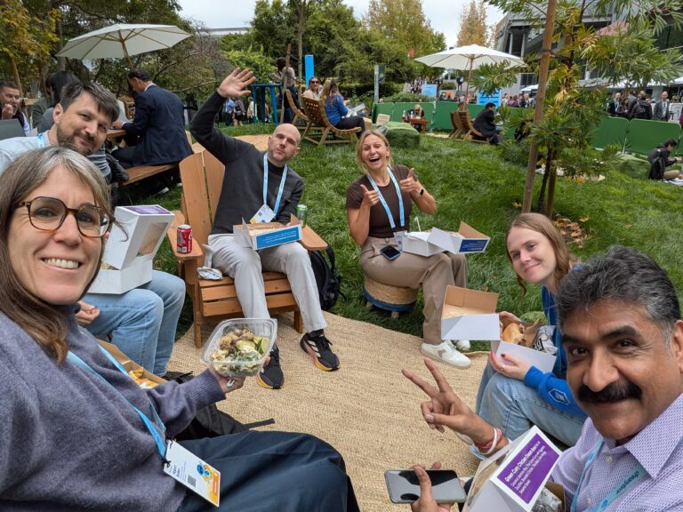 A group of six people sitting outdoors on wooden chairs, eating boxed lunches, with conference badges visible. There are other people and umbrellas in the grassy background.