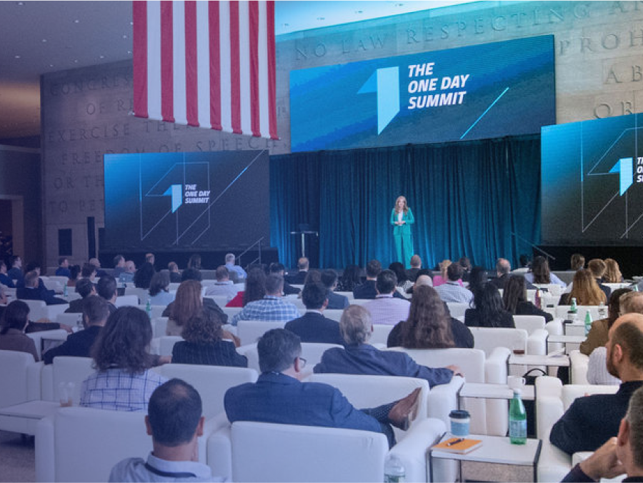 A speaker presents on stage at "The One Day Summit" to an audience seated in a modern conference hall with an American flag overhead.