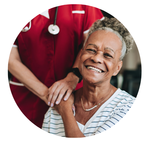 Smiling older woman sits while a healthcare worker in a red uniform stands behind her, resting hands on her shoulders.