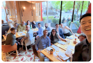 A group of people sit around a long dining table in a restaurant, smiling at the camera. Large windows reveal a city street and greenery outside.