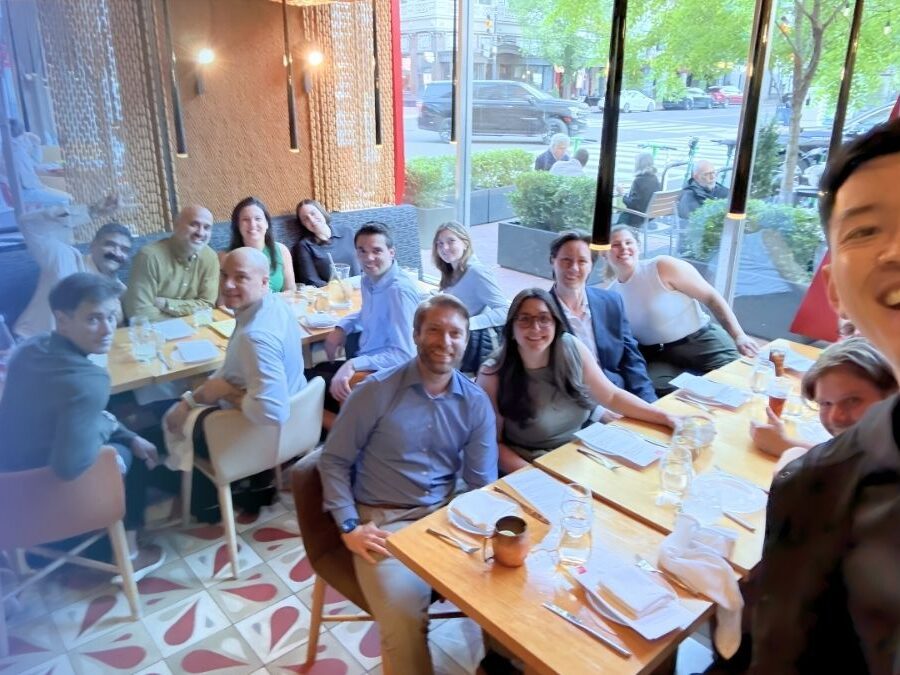 A group of people pose for a photo around two tables set for a meal in a restaurant with large windows showing a street outside.