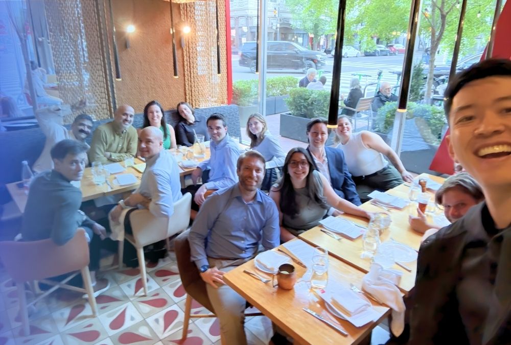 A group of people pose for a photo around two tables set for a meal in a restaurant with large windows showing a street outside.
