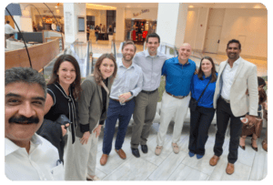 A group of eight adults standing together indoors, smiling at the camera in a shopping mall or office atrium.