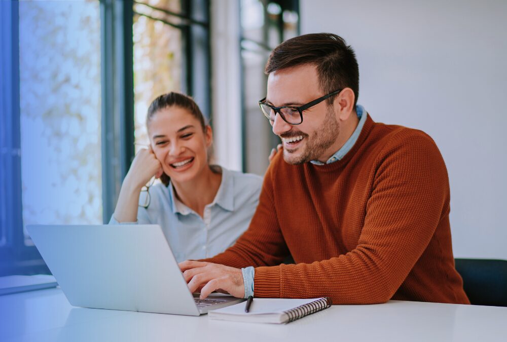 Two people sitting at a desk, smiling and looking at a laptop screen with a notebook and pen nearby in a bright office setting.