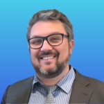 Smiling man with glasses, beard, and mustache wearing a dark suit, striped shirt, and patterned tie, posing in front of a blue gradient background.