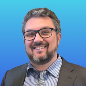 Smiling man with glasses, beard, and mustache wearing a dark suit, striped shirt, and patterned tie, posing in front of a blue gradient background.