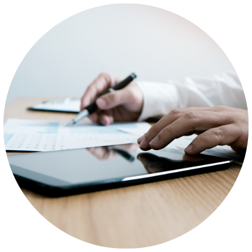 A person reviews printed charts and writes with a pen, while a tablet lies on a wooden desk nearby.