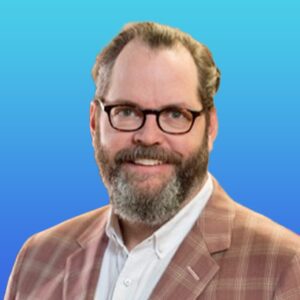 A man with glasses, a beard, and mustache is wearing a plaid jacket and white shirt, smiling at the camera against a blue gradient background.