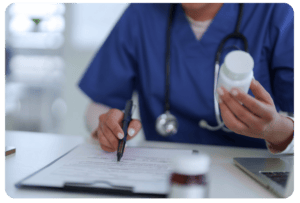 A healthcare professional in blue scrubs holds a medication bottle and writes on a clipboard at a desk with a laptop and medicine containers.