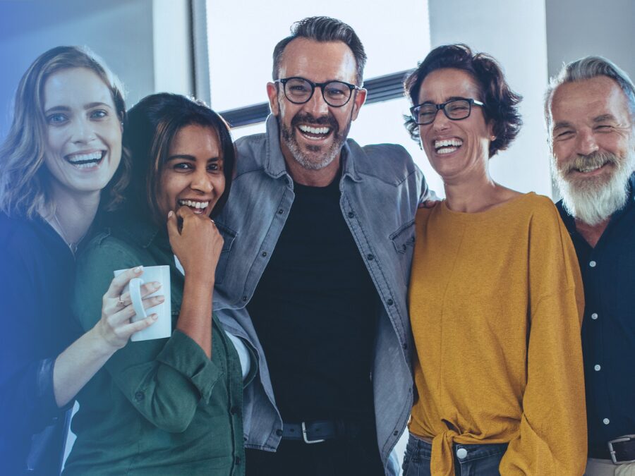 Five adults standing close together indoors, smiling and laughing, with arms around each other.