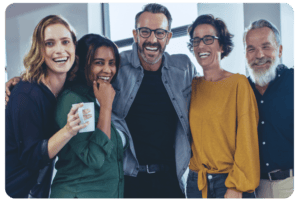 Five adults stand close together indoors, smiling and laughing. One person holds a white mug. The group’s body language suggests camaraderie in a casual or professional setting.