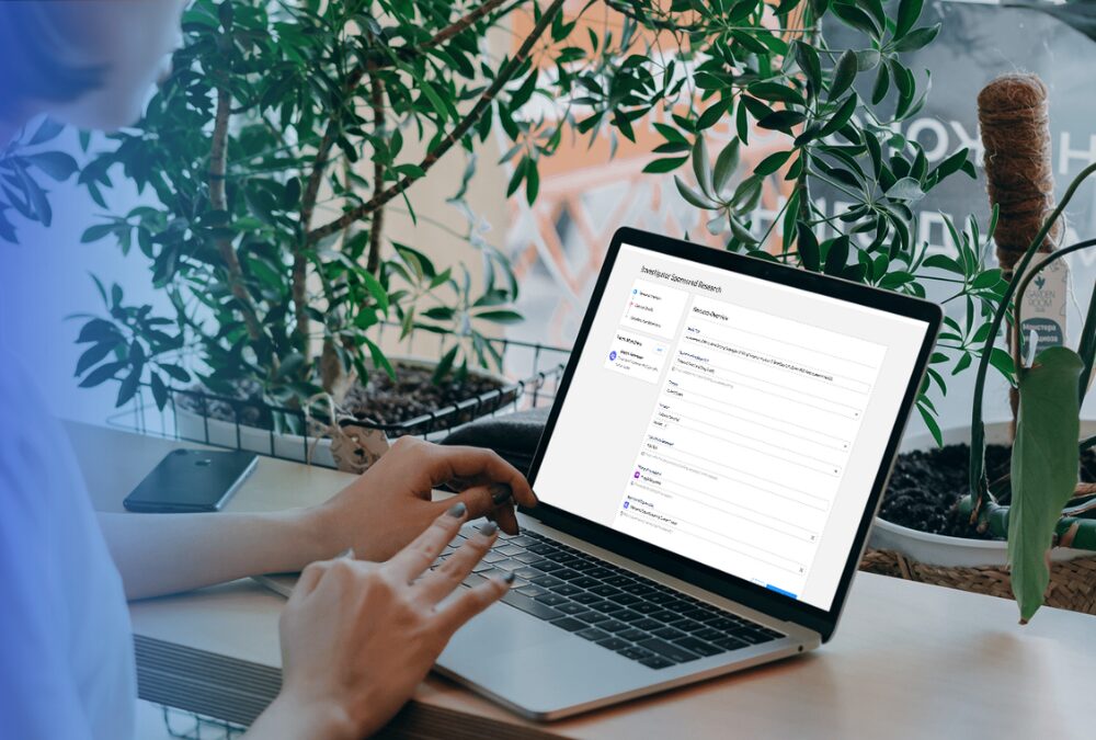Person typing on a laptop with a form titled “Investor Data Questionnaire” on the screen, sitting at a wooden desk near green plants.