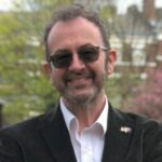 A man with short hair, glasses, and a beard is smiling outside. He is wearing a dark blazer, white shirt, and a small pin on his lapel. The background shows trees and buildings.