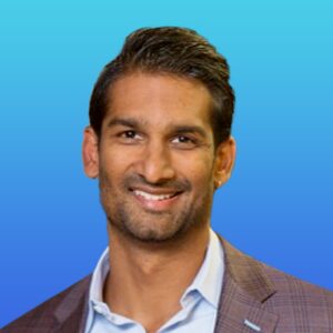 Smiling man with short dark hair wearing a checked suit jacket and light blue shirt, posed in front of a blue gradient background.