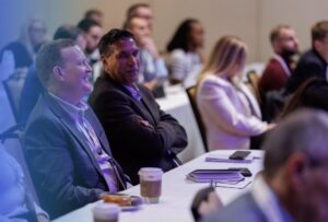 People sit at tables with coffee cups and notepads, attentively listening at a conference or seminar in a professional setting.