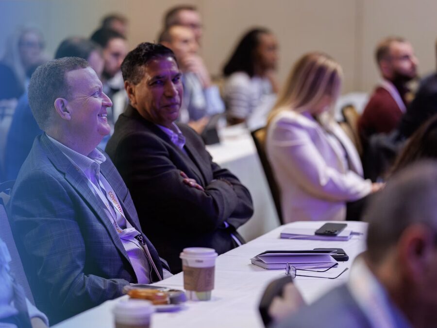 People sit at tables with coffee cups and notepads, attentively listening at a conference or seminar in a professional setting.