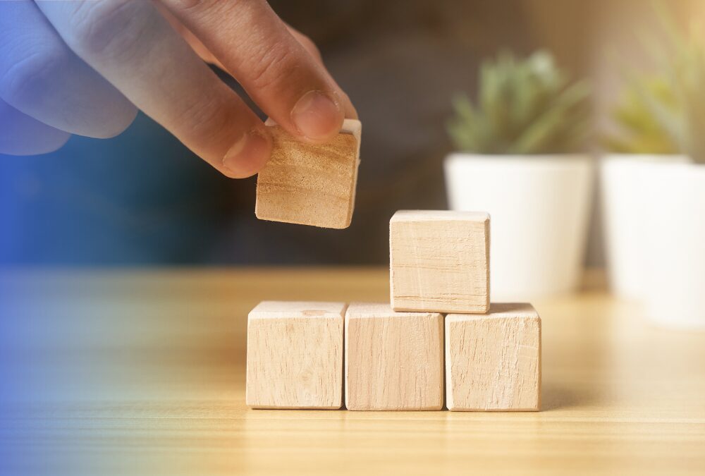 A hand places a wooden block on top of three other wooden blocks on a wooden surface, with two potted plants blurred in the background.
