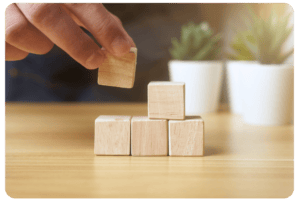 A hand places a wooden block on top of three blocks stacked on a wooden table, with two potted plants blurred in the background.
