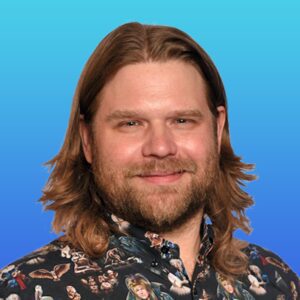 A man with shoulder-length brown hair and a beard, wearing a patterned shirt against a blue gradient background.