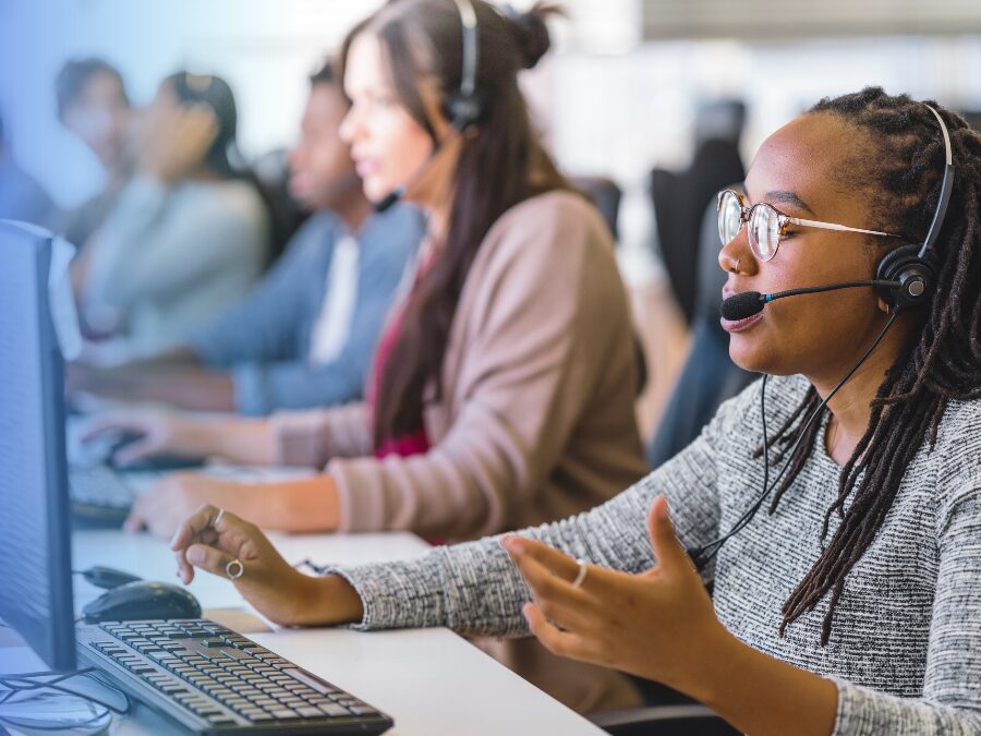 A woman with glasses and a headset speaks while working at a computer in a call center, with other employees working in the background.