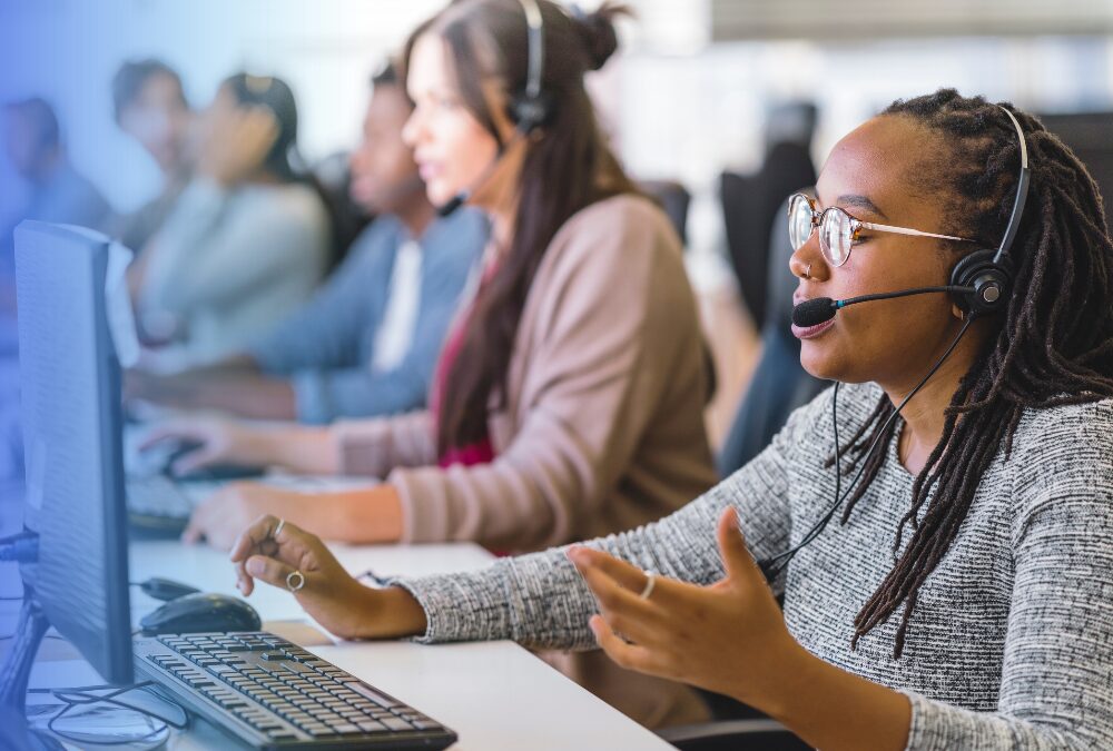 A woman with glasses and a headset speaks while working at a computer in a call center, with other employees working in the background.