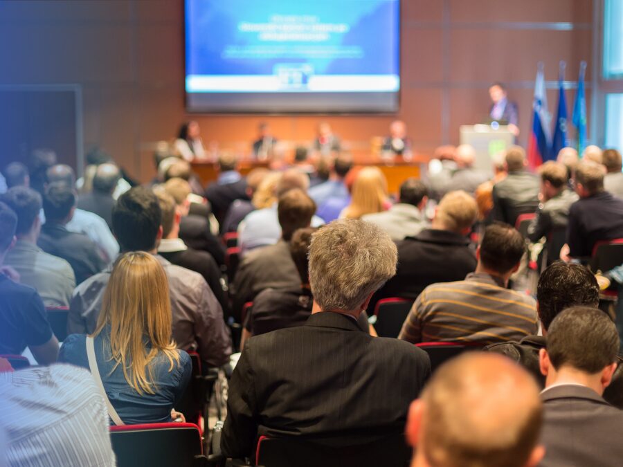 Audience seated in a conference room listening to a panel of speakers at the front, with a presentation projected on a screen.