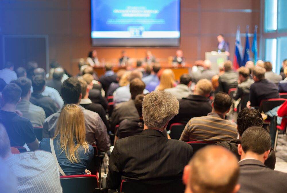 Audience seated in a conference room listening to a panel of speakers at the front, with a presentation projected on a screen.